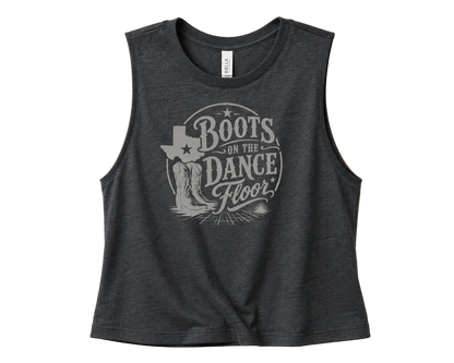 A dark heather gray sleeveless crop top with a distressed light gray graphic reading 'Boots on the Dance Floor' and a Texas-shaped boot design, shown on a solid beige background.
