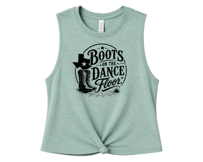 A sleeveless heathered mint green crop top with a black graphic print reading 'Boots on the Dance Floor' featuring cowboy boots and a Texas state silhouette, styled with a front knot detail against a solid peach background.