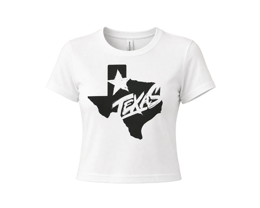A white short-sleeve crop top t-shirt featuring a black graphic of the state of Texas with a white star and the word 'TEXAS' in stylized lettering, displayed on a solid beige background.